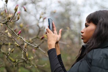 Genç Afrikalı kız öğrenci parkta bahar çiçeklerinin telefonunda fotoğraf çekiyor..