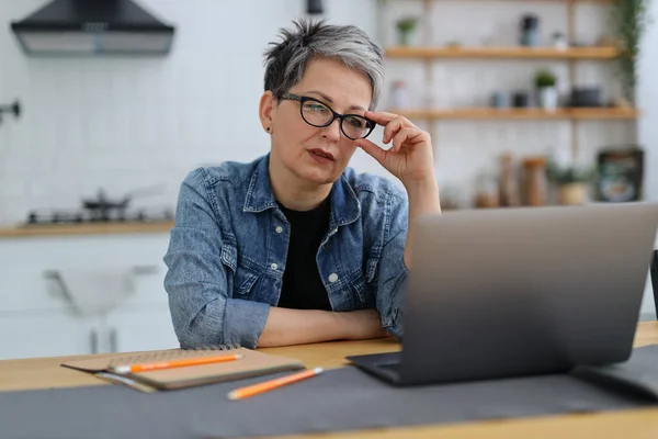 Concentrated mature woman thinks at the workplace, holds glasses with her hand and looks into a laptop.