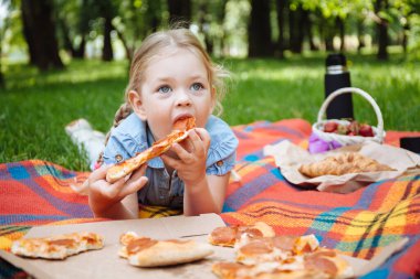 Küçük bebek pizza yiyor ve arka planda yeşil çimenlerin üzerinde gülüyor, yaz pikniği. Parkta dinlen..