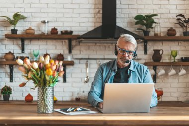 Evden uzak bir iş. Gri saçlı ve sakallı olgun bir adam mutfaktaki ofiste iş görüşmeleri yapıyor. Çevrimiçi video konferansı.