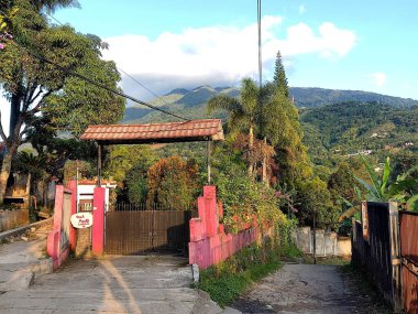 Evin girişi kırmızı çitli ve çatısı olan yeşil ağaçlarla çevrili, arka planda güzel bir dağ manzaralı..