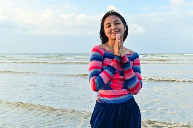 Çizgili elbiseli Asyalı genç kadın gülümsüyor ve parmaklarını denizin önündeki kameraya gösteriyor, mavi gökyüzü arka planında.
