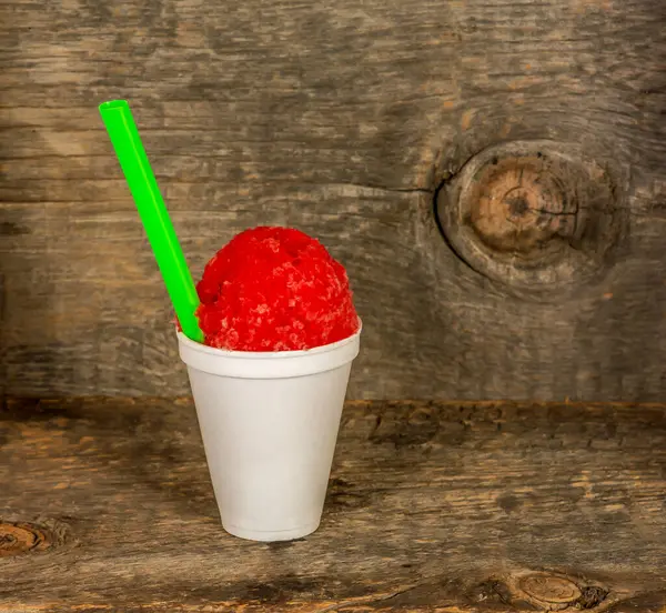 Cherry Red Hawaiian Shave ice, shaved ice or snow cone dessert in a white cup with a green straw on a rustic barn wood background.