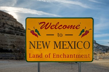 New Mexico yol tabelasına hoş geldiniz. Arkasında bulutlu bir gökyüzü ve dağlar var..