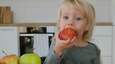 Çocuk mutfakta kırmızı bir elma ısırıyor.