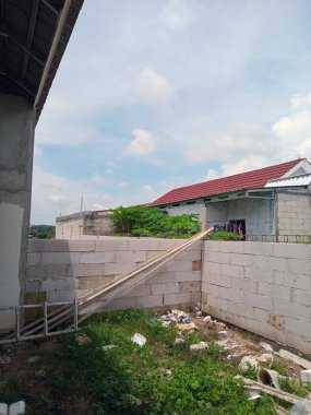 Beton duvarlı, çimenli ve dağınık enkazlı, bakımsız bir açık hava manzarası. Arka planda, kırmızı çatılı ve kıyafetleri dışarıda asılı iki bina var.