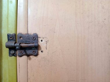 Ahşap kapıdaki paslı kapı menteşesinin yakın çekim görüntüsü. Menteşe aşınma ve yaşlanma belirtileri gösteriyor. Açık renkli ahşap bir yüzey ve kısmen görülebilir bir mandal..