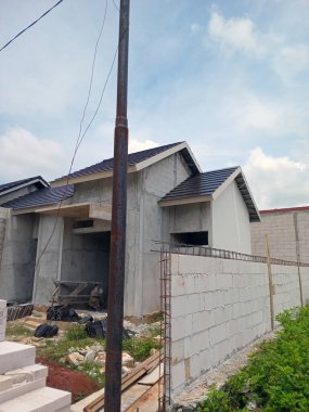 Kısmen inşa edilmiş beton yapısı ve eğimli çatısı olan bir ev. Bina inşaat malzemeleri ve döküntülerle çevrili ve arka planda açık mavi bir gökyüzü var..