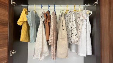 Ahşap bir gardıroptaki renkli askılarda asılı çocuk kıyafetleri..