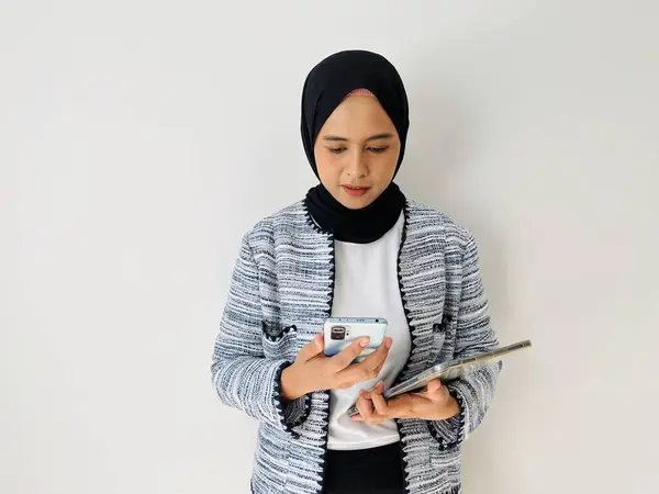 stock image Young Asian woman smiling while using mobile phone. Standing female wearing black hijab and tweed blazer isolated on white background.