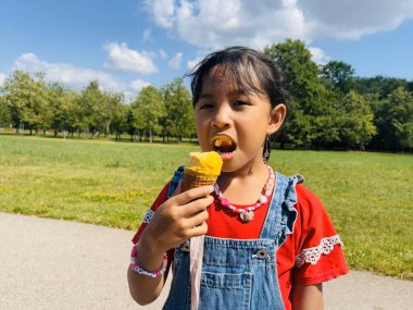 Beş yaşındaki Asyalı bir çocuk yaz mevsiminde dışarısı sıcakken dondurma yer. Waffle külahında mango dondurması. Parkta mutlu ve mutlu bir çocuk.