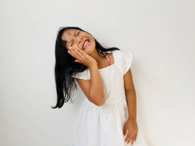 Beş yaşında Asyalı bir kız, bir eli yanağında, gözleri kapalı gülümsüyor.