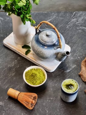 Seramik bardakta kibrit sütlü latte, parlak yeşil kibrit tozu ve seramik çaydanlığın yanında. Modern ve minimalist bir ortam. Sağlık ve yaşam tarzı içeriği, çay blogları ve kafe menüleri için.