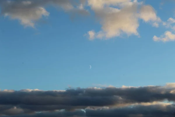 Moon between the clouds in the blue sky, birds flying in the distance, dark clouds below