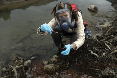 Gaz maskeli ve koruyucu lateks mavi eldivenli biyolog kurumuş bir yapay göl yatağından Pincette ile toprak örnekleri alıyor..