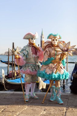 Venedik, Veneto, İtalya - 19 Şubat 2020: Pastel Renkleri 'nde Zarif Venedik Maskeli Çifti San Marco Meydanı' nın rıhtımında Gondolas Jetty ve San Giorgio Maggiore Kilisesi 'nde poz veriyor.