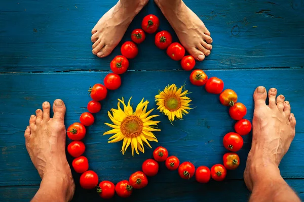 Male and Female Feet around Red Tomatoes forming a Heart Shape on Blue Wooden Table Background  with two Sunflowers inside the Heart - Love for Organic Food and Healthy Lifestyle Concept