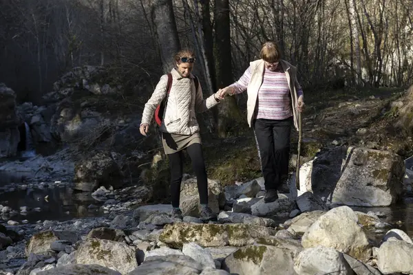Helping Senior Woman Walk over a Wild Stream Of Water in Nature While on a Refreshing Walk in Forest Air