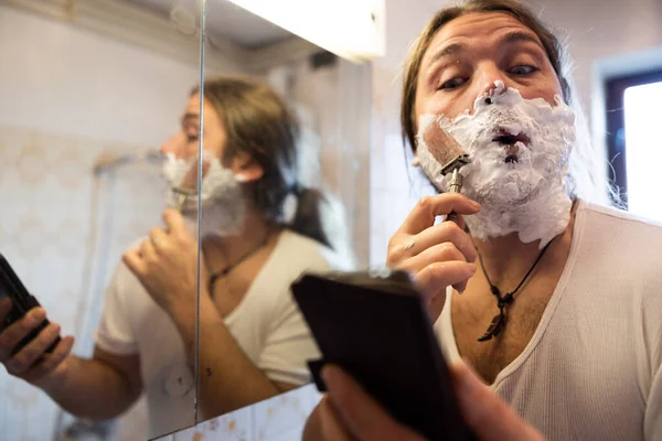 Mid Adult Man Shaving his Beard with help of a Phone instead of the Mirror in Domestic Bathroom Close Up