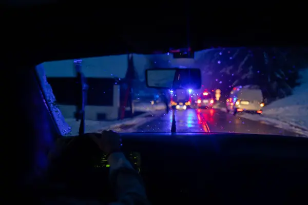 View Through Car Windshield Reveals a Traffic Accident Scene with Flashing Blue Police and Red First Aid Ambulance Lights, Capturing the Urgency and Challenges of Slippery Winter Roads