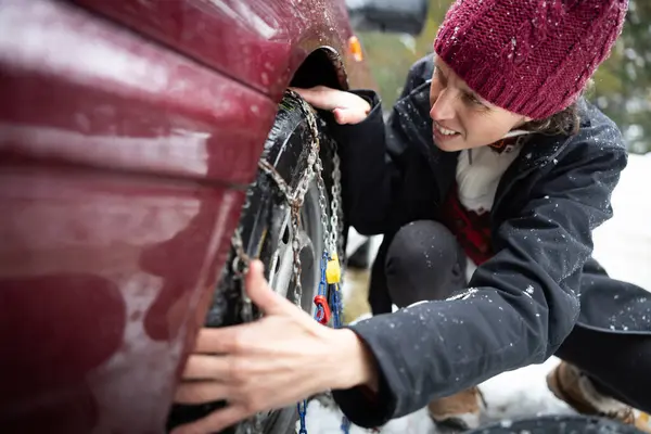 Determined Adult Female Driver Faces Icy Cold Weather Challenges, Installing Tire Chains for Snow-Covered, Slippery Roads Amidst a Snowfall
