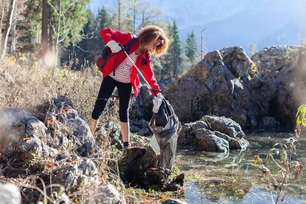 Woman Backpacker Ecologist Picking Up Trash and garbage in nature Environment of a Beautiful Mountain Lake