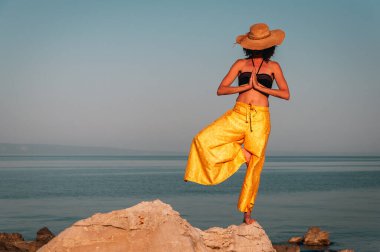 Altın sarısı pantolonlu Kıvırcık Kıvırcık Kadın Bikini ve Hasır Şapka Meditasyon Anları Günbatımında Sıcak Sahilde