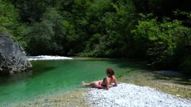 Bikini 'de bir Dağ Nehri plajında uzanan ve İHA' nın Slovenya, Avrupa Alpleri 4K Video 'sundan ferahlatıcı suyun tadını çıkaran kadın.