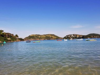 Buzios, Brezilya: Sakin deniz, renkli tekneler, yeşil tepeler ve açık mavi gökyüzü altındaki binalarla sakin sahil sahnesi