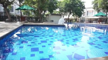 Derin mavi mozaik desenli, etrafındaki ağaç ve binaları yansıtan huzurlu bir havuz sahnesi.