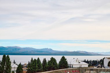 Ağaçlardan oluşan bir çalılıkta, bir gölün ve dağların arkasındaki binalar.