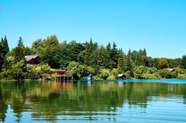 Mavi bir göl yakınında yeşil bitki örtüsü olan kıyı, ufukta dağlardan oluşan bir manzara ve orman içinde evleri olan Bariloche Ulusal Parkı.