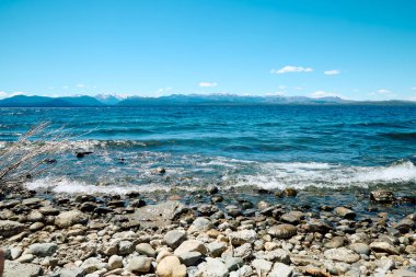Mavi gölün kayalık kıyısı, ufukta karla kaplı dağlardan oluşan bir manzara, Bariloche Ulusal Parkı.
