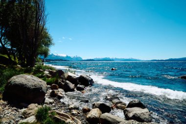 Mavi bir göl yakınında yeşil bitki örtüsü olan kayalık kıyılar ufukta karla kaplı dağlardan oluşan bir manzara, Bariloche Ulusal Parkı