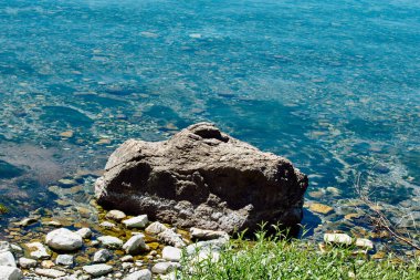 Taşların arasında kayalık kıyı ve yeşil çimen. Şeffaf turkuaz su kıyıları siler..