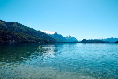 Güzel dağ gölü. Şeffaf mavi su. Etrafı kayalık dağlar ve ormanlarla çevrili. Yüzücülerle uzakta bir tekne..