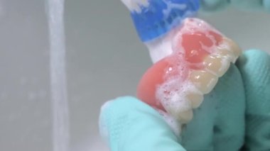 Yapay dişleri fırçayla temizlerken yakın çekim. Her şeyin temiz olduğundan ve lastik eldiven kullandığından emin oluyorum. Arka planda akan su.