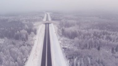 Drone shot of a highway and the snowy forest surrounding it in Estonia. Cold scandinavian winter. Vehicles driving. Cloudy weather. Drone moving backwards and upwards.