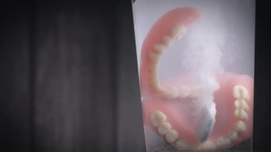 Protez diş temizleyicisi tabletinin suda yavaşça çözünüşünü yakından çek. Bir bardak suda protez diş. Arka planda bulanık tahta var. Boşluğu kopyala.