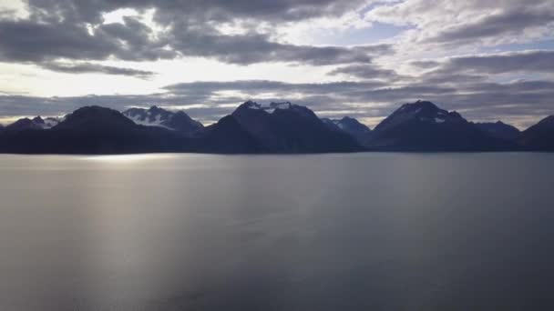 ノルウェーのフィヨルド山の午後の空中観察 海面の太陽を反射する 氷河の氷で覆われた山頂 空は半分曇っている カメラがゆっくり上に移動する — ストック動画