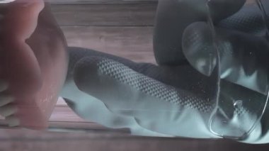 Plastik eldiven kullanılarak bir bardak sudan çıkarılan protez dişlerin yakın çekimi. Arka planda tahta tahta var. Yandan ve aşağıdan yukarıya bak. Dikey Biçim Videosu