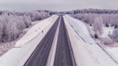 Estonya 'da soğuk bir kış sırasında yüksek açılı bir otoyol manzarası. Hava bulutlu. Araba sürüyor. Karlı orman ve toprak. İnsansız hava aracı yavaşça ilerliyor.