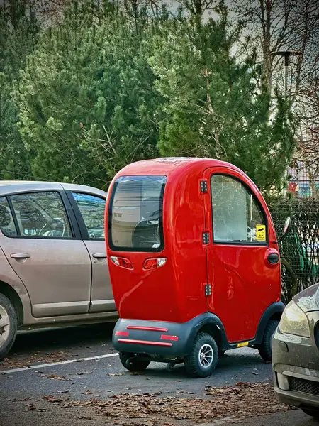 Yuvarlak çatılı kırmızı bir retro mobiletli scooter, ağaçların ve çalıların arasına kurulmuş, modern ve klasik unsurları pürüzsüz bir şekilde harmanlayan gümüş bir arabanın yanına park edilmiştir..