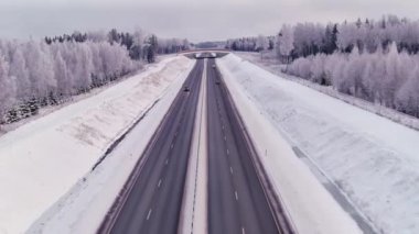 Estonya 'daki soğuk İskandinav kışında yüksek açılı bir otoyol manzarası. Hava bulutlu. Karlı orman ve toprak. Araba sürüyor. İnsansız hava aracı yavaşça ilerliyor.