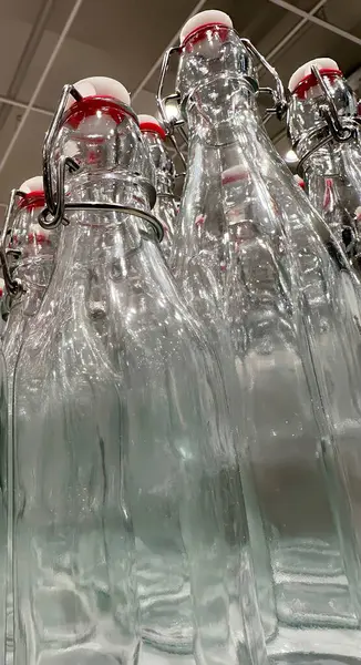 Kırmızı şapkalı şeffaf cam şişelerden oluşan bir kolleksiyon, öğleden sonra ışığı altında eşsiz şekillerini ve yansımalarını sergiledikleri parlak bir mağaza koridorunda yakın bir şekilde düzenlenir..