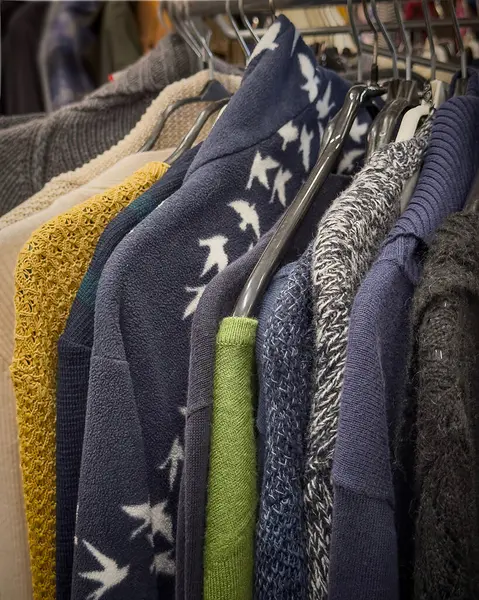 Bir ucuzluk mağazasındaki askılarda çeşitli renkli kazaklar sergilenerek, modanın yeniden kullanımı ve geri dönüşümü kavramları tanıtılıyor. Müşteriler sürdürülebilir giyim seçeneklerini gözden geçirirler.