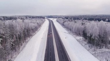 Kışın Estonya 'da dört şeritli bir otoyolun hava görüntüleri. Bulutlu gökyüzü. Karlı orman ve toprak. Soğuk İskandinav havası. Araçlar sürüyor. Drone yavaşça uzaklaşıyor.