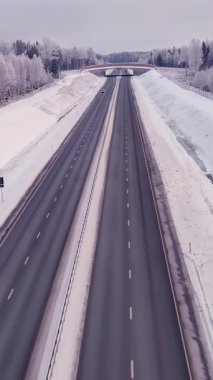 Estonya 'da soğuk bir kış sırasında yüksek açılı bir otoyol manzarası. Hava bulutlu. Araba sürüyor. Karlı orman ve toprak. Drone yavaşça ilerliyor. Dikey video
