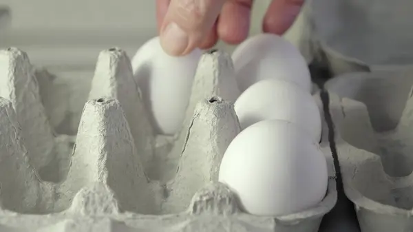 Yumurtaları bir eliyle bir yumurtalıktan diğerine taşıyor. Bir sürü büyük beyaz yumurta var. Arka plan bulanık. Yakın çekim..