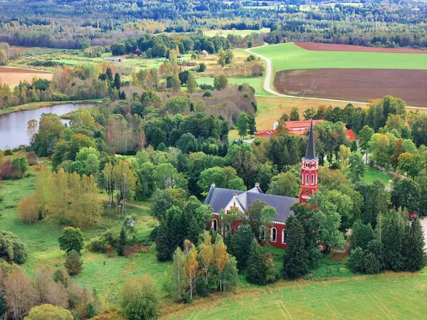 Halliste, Estonya 25 Eylül 2025: Kızıl kilise yeşil sonbahar ağaçlarına kurulmuş, tarlalarla çevrili, küçük bir göl ve kırsal Estonya kırsal kesimi yukarıdan görülüyor
